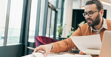 man looking at a document with a pen in hand next to a laptop